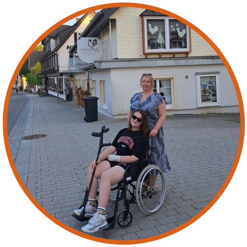 An image of Phoebe, a CRPS patient, seated in a wheelchair and wearing sunglasses and a black t-shirt, alongside Rachel, her mother, standing behind her with a hand on the wheelchair. They are on a quiet street with quaint buildings in the background.