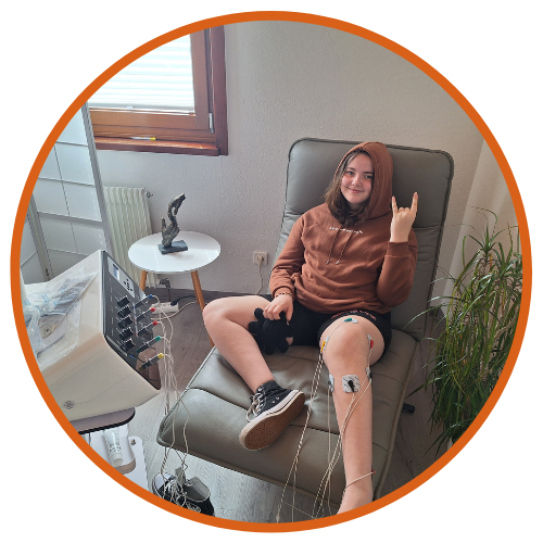 Phoebe sits relaxed in a reclining chair, offering a "rock on!" hand gesture, while undergoing Scrambler Therapy treatment. Leads from the therapy machine are attached to her leg, indicating the non-invasive nature of the procedure. The room is bright, with a side table and plant, creating a calm therapeutic environment.