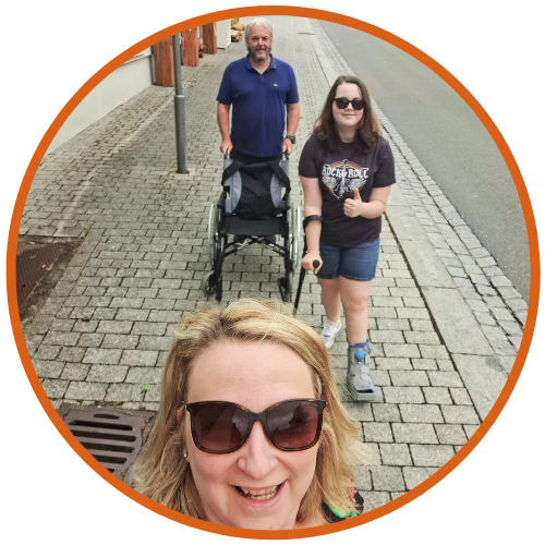 A celebratory moment captured in a circular-framed photo with Rachel in the foreground and Phoebe walking behind her. Mick, her father, is pushing an empty wheelchair in the background, signifying the family's progress. They are on a pedestrian street that underscores the family's joyous occasion.