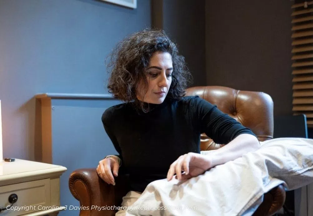 A sitting woman looks tentatively at her arm placed on a cushion. She is sat in a physiotherapy treatment room.