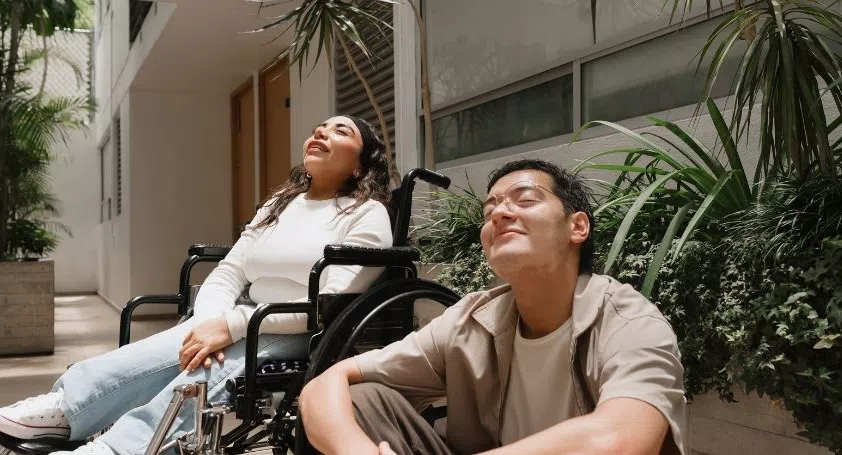 A girl sitting in a wheelchair and a man sitting on the stairs, both with their heads tilted up and eyes closed, enjoying the sunshine