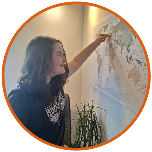 Phoebe, with a smile, is pointing at Germany on a world map, indicating the destination for her Scrambler Therapy treatment. The setting is a well-lit room with a subtle decor and a potted plant to the side.