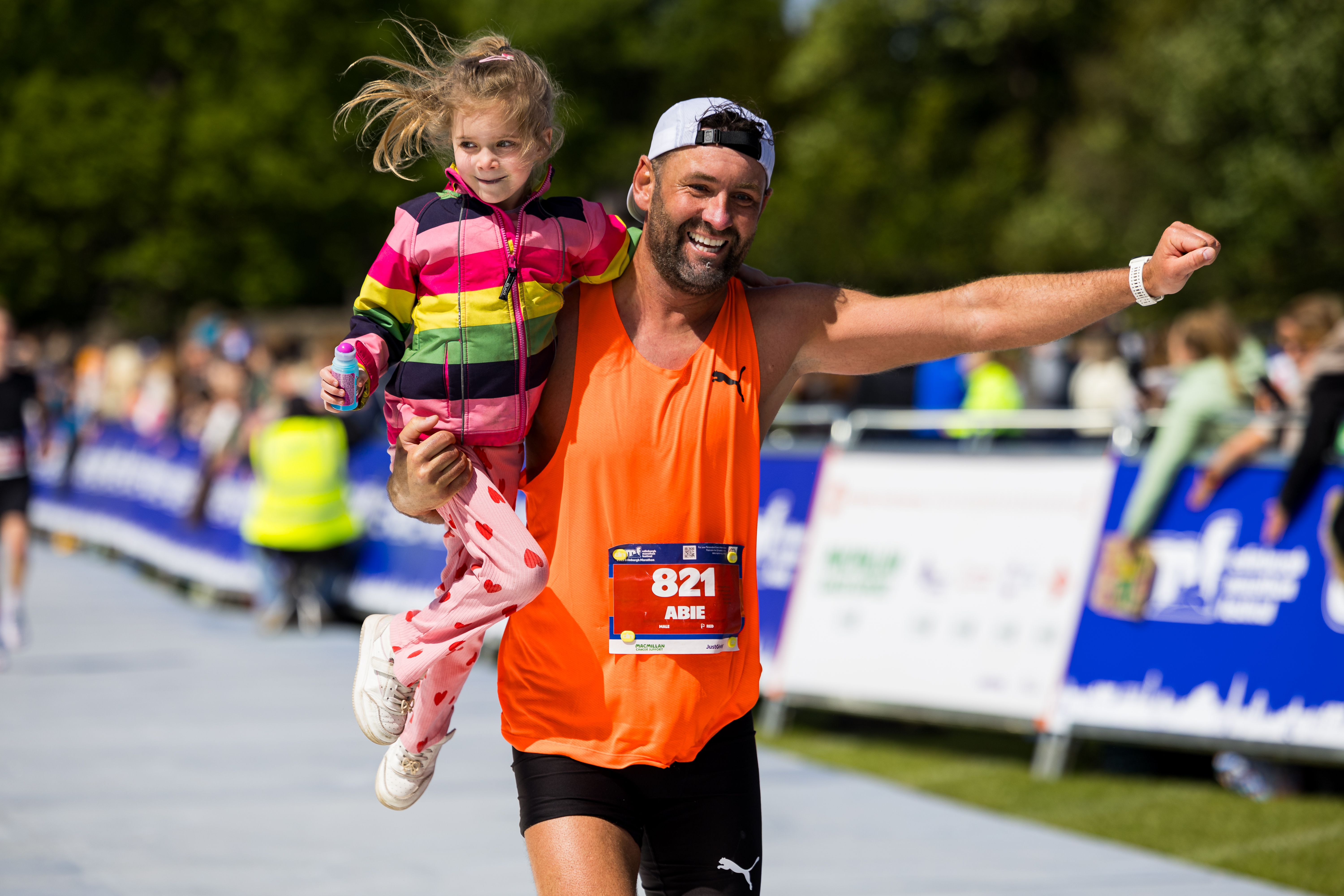 2026 Edinburgh Marathon Festival, photo of event participant in an orange sports vest running along the route, holding a smiling child in one arm and punching the air triumphantly with the other arm