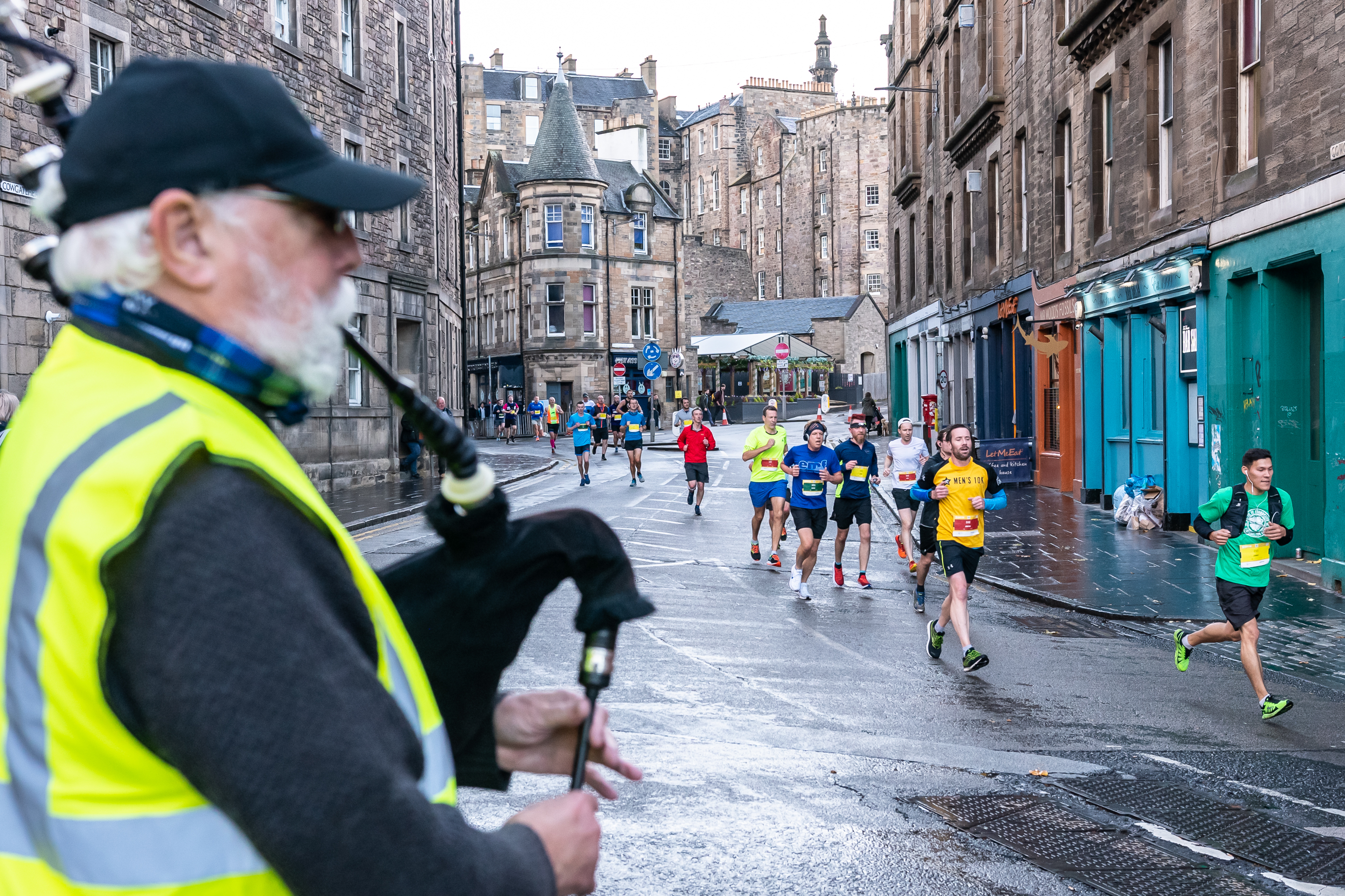 2026 Men’s 10K Edinburgh, photo of event participants running along the route with a man in a high-vis vest playing the bagpipes in the foreground