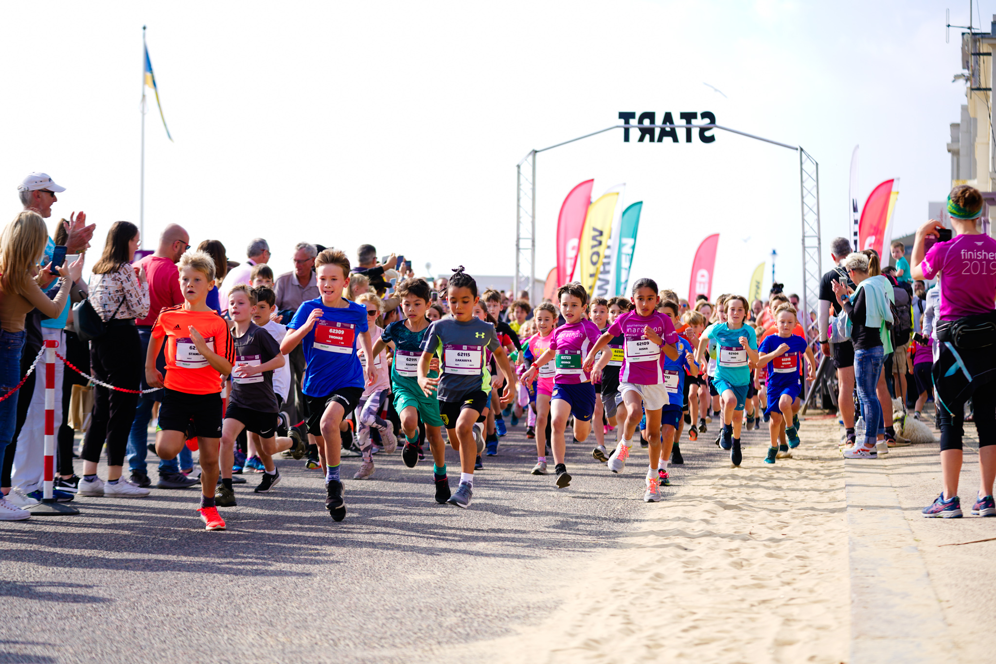 2026 Run Bournemouth Weekend, photo of children crossing the start line for one of the races for young people, there are crowds cheering on each side and the start line is in the background as the children run along a sandy path towards the camera