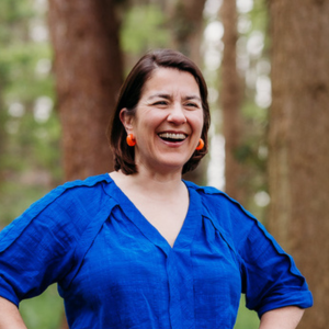 Laughter Yoga, photo of Elspeth Fimpel laughing with a blurred woodland scene in the background, Elspeth is wearing a vibrant blue linen blouse and orange earrings and she has short brunette hair