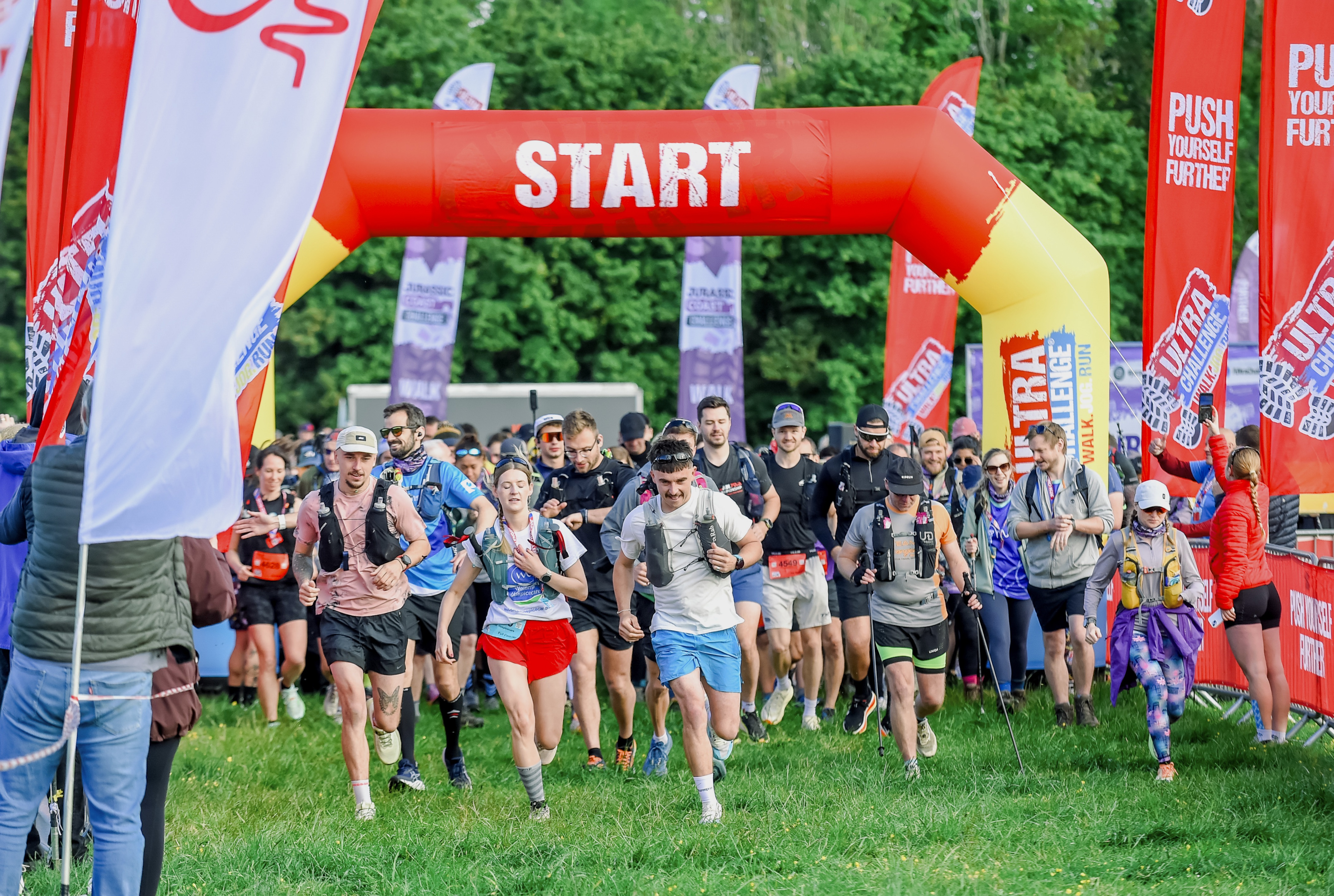 Image of a wave of runners crossing the start line at an event, there are big banners and crowds cheering on both sides as they run towards the camera
